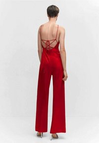 Une combinaison rouge présente une finition satinée, un dos ouvert avec des bretelles croisées, des jambes larges et un buste ajusté. Des talons aiguilles argentés sont visibles.