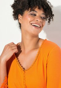 Oranžový žebrovaný top s výstřihem do V a dekorativním květinovým krajkovým lemem kolem výstřihu a rukávů. Modelka se usmívá s přirozenými kudrlinami.