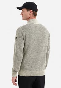 Man die zijwaarts kijkt en een gestructureerde lichtgrijze gebreide trui en een zwarte baseballcap draagt, tegen een witte achtergrond.