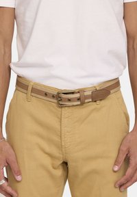 Bruine leren riem met een gestructureerde afwerking, voorzien van een metalen gesp en contrasterende beige stiksels, gedragen over kaki broek.