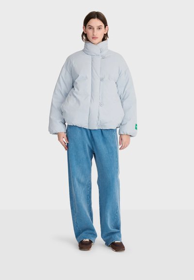 Femme portant une doudoune gris clair avec col montant et pantalon large bleu, debout devant un fond blanc uni.