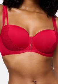 Sustentação vermelha com copas almofadadas e acabamentos em renda. Apresenta alças ajustáveis e um detalhe central em tom dourado na parte da frente. Textura suave.