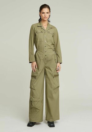 Femme debout portant une combinaison utilitaire à manches longues vert olive avec poches cargo et bottes noires à bout carré sur un fond uni.