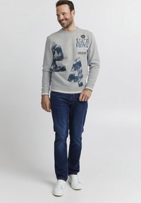 Grauer Pullover mit blauem Grafikdruck, der die Zahl "24" zeigt, kombiniert mit dunkelblauen Jeans und weißen Sneakern. Einfaches Design, lässige Passform.
