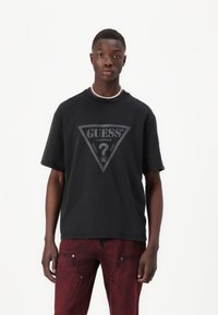 Melna kokvilnas T-kreklā ar lielu pelēku trīsstūra logotipu, kurā redzams "GUESS" un "ORIGINALS." Veidots ar īsām piedurknēm un apaļu kakla izgriezumu.