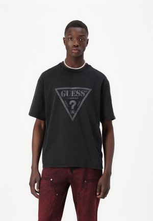 T-shirt in cotone nero con un grande logo triangolare grigio che presenta la scritta "GUESS" e "ORIGINALS". Design con maniche corte e scollo rotondo.