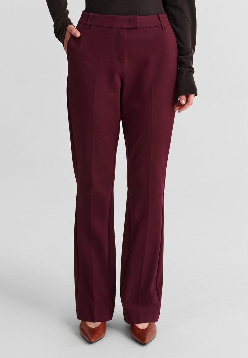 Femme portant un pantalon bordeaux taille haute à jambes droites avec poches latérales, associé à des chaussures marron à bout pointu et un haut noir à manches longues.