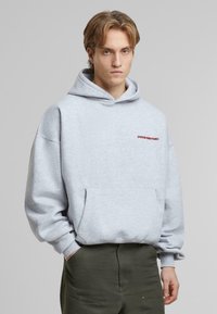 Lysegrå hettegenser med frontlomme, løs passform og snor i hetta. Har "FORBUDT" i rød brodering på brystet.