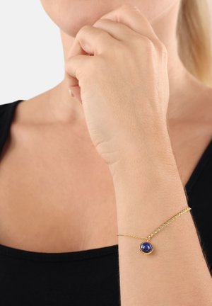 Primer plano de una persona que lleva una camiseta negra y una pulsera dorada con una gema azul en su muñeca.