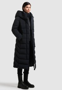 Kvinde iført lang sort quiltet dunjakke med hætte og sorte støvler, stående mod en ensartet lys baggrund.