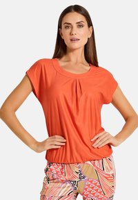 Kurzärmliges oranges Oberteil aus glattem Stoff, mit rundem Ausschnitt und einer Faltennaht vorne, kombiniert mit gemusterten Hosen.
