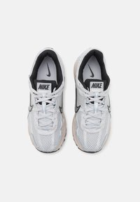 Tênis Nike com um upper de malha respirável em cinza claro, detalhes em preto, atacadores planos e uma sola acolchoada em bege.