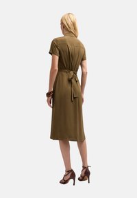 Rochie midi verde olive cu mâneci scurte, talie ajustată și detaliu cu fundiță la spate, confecționată dintr-un material neted. Asortată cu tocuri maro.