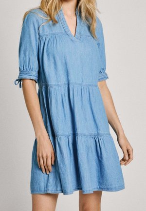 Femme portant une robe courte en jean bleu clair avec jupe à volants et manches au coude nouées aux poignets, debout devant un fond uni.