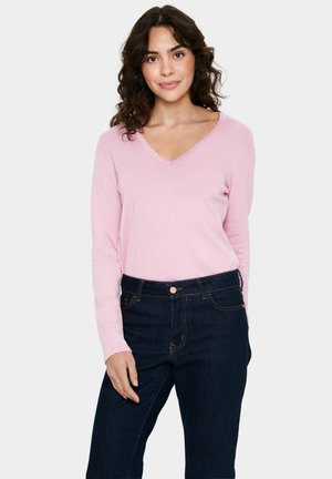 Lichtroze lange mouwen V-hals top met golvende randen, gecombineerd met donkerblauwe high-waisted jeans. De stof lijkt zacht en gestructureerd.