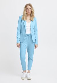 Vrouw met blond haar in een lichtblauwe blazer en bijpassende elastische broek, witte tanktop en witte sneakers, staande tegen een effen achtergrond.