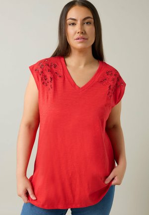 Kvinde med langt brunt hår iført en rød ærmeløs top med V-hals og sorte broderede blomsterdesign på skuldrene, stående mod en lys baggrund.