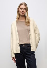 Krämsfärgad cardigan i mjukt, strukturerat tyg, med avslappnad passform, knappfront och långa ärmar, bärs över en beige topp.