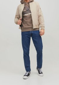 Chaqueta bomber de ante beige sobre una sudadera marrón, combinada con jeans de mezclilla azul y zapatillas negras. La sudadera presenta un estampado de logo.