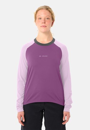 Jong persoon met licht krullend haar draagt een paars en lichtroze shirt met lange mouwen en een zwarte broek, staand tegen een effen achtergrond.