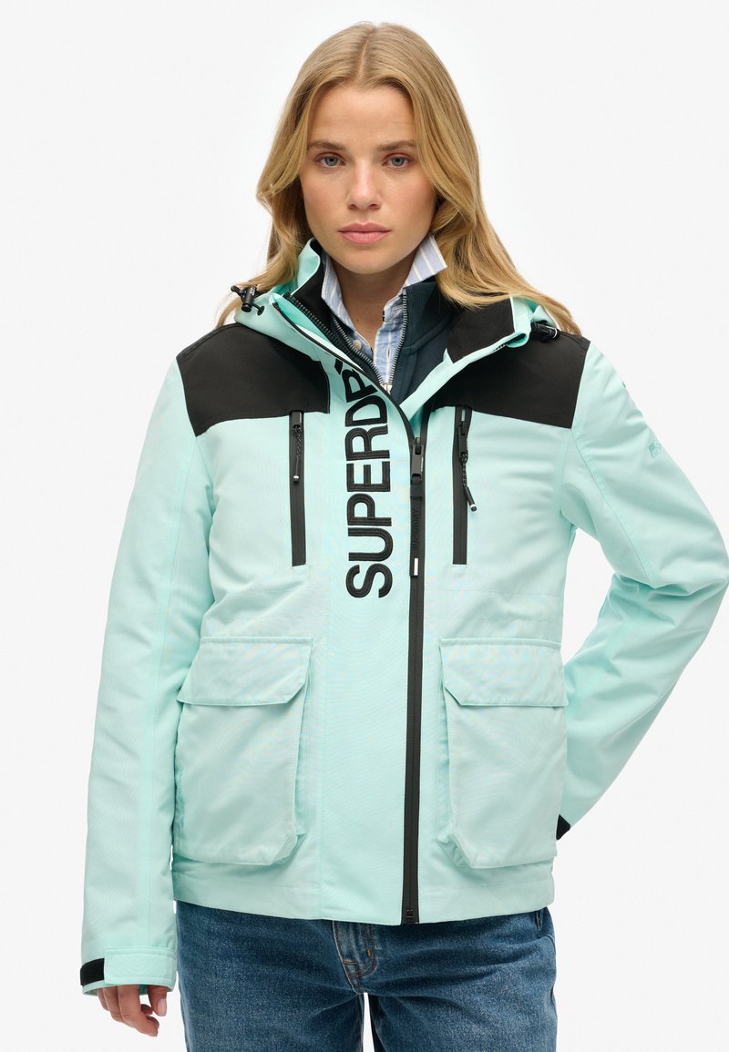 Lichtblauwe jas met zwarte accenten, met ritszakken en een prominent logo. Heeft een capuchon en een gestructureerde afwerking. Geschikt voor buitengebruik.