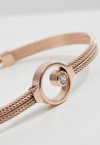 Bracelet en métal rose doré avec une texture tressée, présentant un accent circulaire et une pierre claire au centre. Design élégant et moderne.
