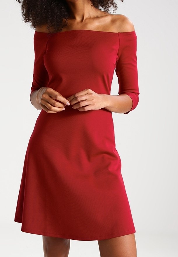 Femme portant une robe rouge ajustée, longueur genou, décolletée, avec des manches trois-quarts, debout devant un fond clair uni.