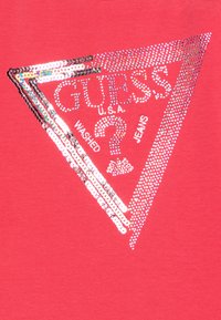 Koralo raudonos audinio paviršius turi trikampį raštą, kuris išsiuvinėtas sidabro blizgučiais, užrašant "GUESS U.S.A. WASHED JEANS" ir klausimo ženklą viduryje.