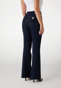 Dunkelblaue Jeans mit weitem Bein und hohem Bund, ausgestattet mit Gesäßtaschen und dekorativen Knöpfen an der Seite. Glatte Textur, klassisches Design.