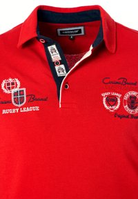 Rød polo-shirt lavet af bomuld, med en marineblå krave, broderede logoer og en to-knaps placket med kontrastdetaljer og syninger.