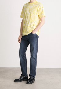 Man som bär en gul mönstrad kortärmad skjorta, mörkblå jeans, svarta läderskor, står med ena handen i fickan mot en vit bakgrund.