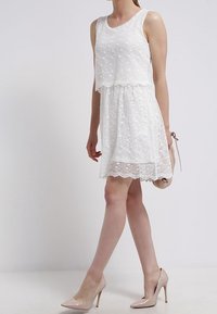 Weißes Spitzenkleid mit ärmellosem Design, gewelltem Saum und geschichtetem Effekt. Getragen mit nuden hohen Schuhen und einer kleinen Handtasche.