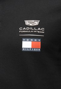 Μαύρο ύφασμα με λευκό λογότυπο της ομάδας Formula 1 της Cadillac πάνω από το ορθογώνιο λογότυπο σημαίας της Tommy Hilfiger σε κόκκινο, λευκό και μπλε.