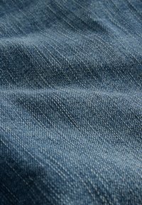 Tejido de denim en tono azul medio, que muestra un tejido texturizado con costuras visibles. La superficie parece suave con un ligero brillo.