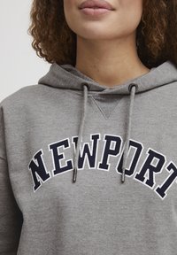 Felpa grigia in cotone con tasca a marsupio frontale, cordini e grafica testuale bianca "NEWPORT" in blu navy. Tessuto testurizzato con una vestibilità casual.