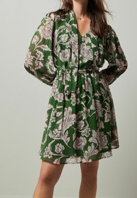 Abito floreale verde in tessuto leggero e trasparente. Caratterizzato da maniche a sbuffo, scollatura a V e vita cin cin cintrata con una cintura di cravatta.