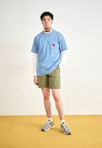 Camiseta azul de manga corta con un bolsillo y un logotipo de corazón rojo, combinada con pantalones cortos verde oliva y zapatillas deportivas grises. Suelo amarillo.
