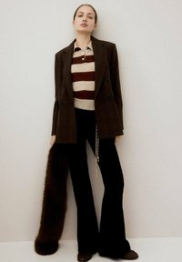 Blazer de cuadros marrón sobre un suéter a rayas crema y burdeos, combinado con pantalones de campana negros y zapatos marrones. Accesorios incluyen una bufanda de piel sintética.