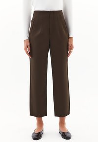 Pantaloni dritti a vita alta marroni indossati con una maglietta bianca a maniche lunghe e scarpe slip-on nere, mostrati su una persona in piedi.