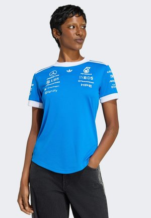 Vrouw die een blauw Mercedes-AMG Petronas raceteamshirt draagt met sponsorlogo's, staand met één hand in de zak en schuin kijkend.