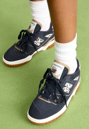 Osoba nosząca czarne buty sportowe New Balance z białymi i beżowymi akcentami oraz białe prążkowane skarpetki typu crew na jasnawo-zielonym dywanie.