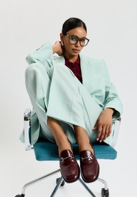 Mátově zelený oblek se širokými nohavicemi, burgundská košile a lesklé burgundské loafers. Průhledné brýle a stříbrné akcenty na náušnicích, sedící na židli.