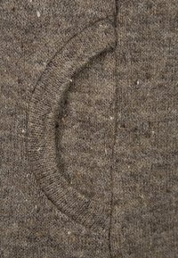 Tessuto lavorato a maglia marrone con una finitura texturizzata, caratterizzato da una tasca curvata cucita nel materiale. Sono visibili delle piccole macchie di colori più chiari.
