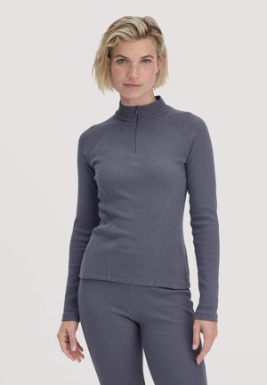 Vrouw met kort blond haar in een aansluitende grijze ribstof long sleeve top met een korte rits en bijpassende broek, staande tegen een witte achtergrond.