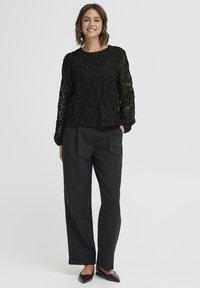 Crna čipkasta majica s složenim cvjetnim uzorcima, okruglim izrezom i dugim rukavima, u kombinaciji s crnim hlačama visokog struka i bordo cipelama.