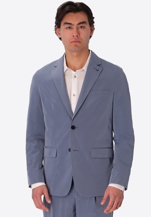 Uomo che indossa una giacca da abito azzurro chiaro sopra una camicia bianca, in piedi davanti a uno sfondo bianco semplice.