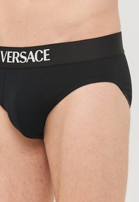 Must muströöv mustrõhv, millel on elastne vöökoht ja valge "VERSACE" bränding. Kujunduses on toetuseks kontuuriga esikott.