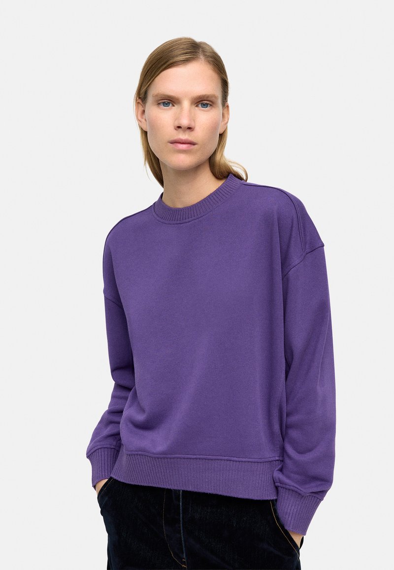 Fioletowa bluza z okrągłym dekoltem, opadającymi ramionami i ściągaczami na mankietach. Gładka faktura, luźny krój i minimalistyczny design.