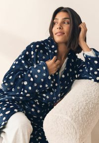 Tamsiai mėlynas flisinis chalatas su baltomis taškeliais, šaliko apykakle ir šoninėmis kišenėmis, dėvimas ant šviesios spalvos aprangos, atsirėmęs į tekstūruotą fotelį.