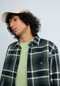 Grünes und schwarzes kariertes Hemd mit weißen Akzenten, mit Knöpfen, über einem hellgrünen T-Shirt getragen. Beiger Hut auf dem Kopf, strukturierte Locken sichtbar.